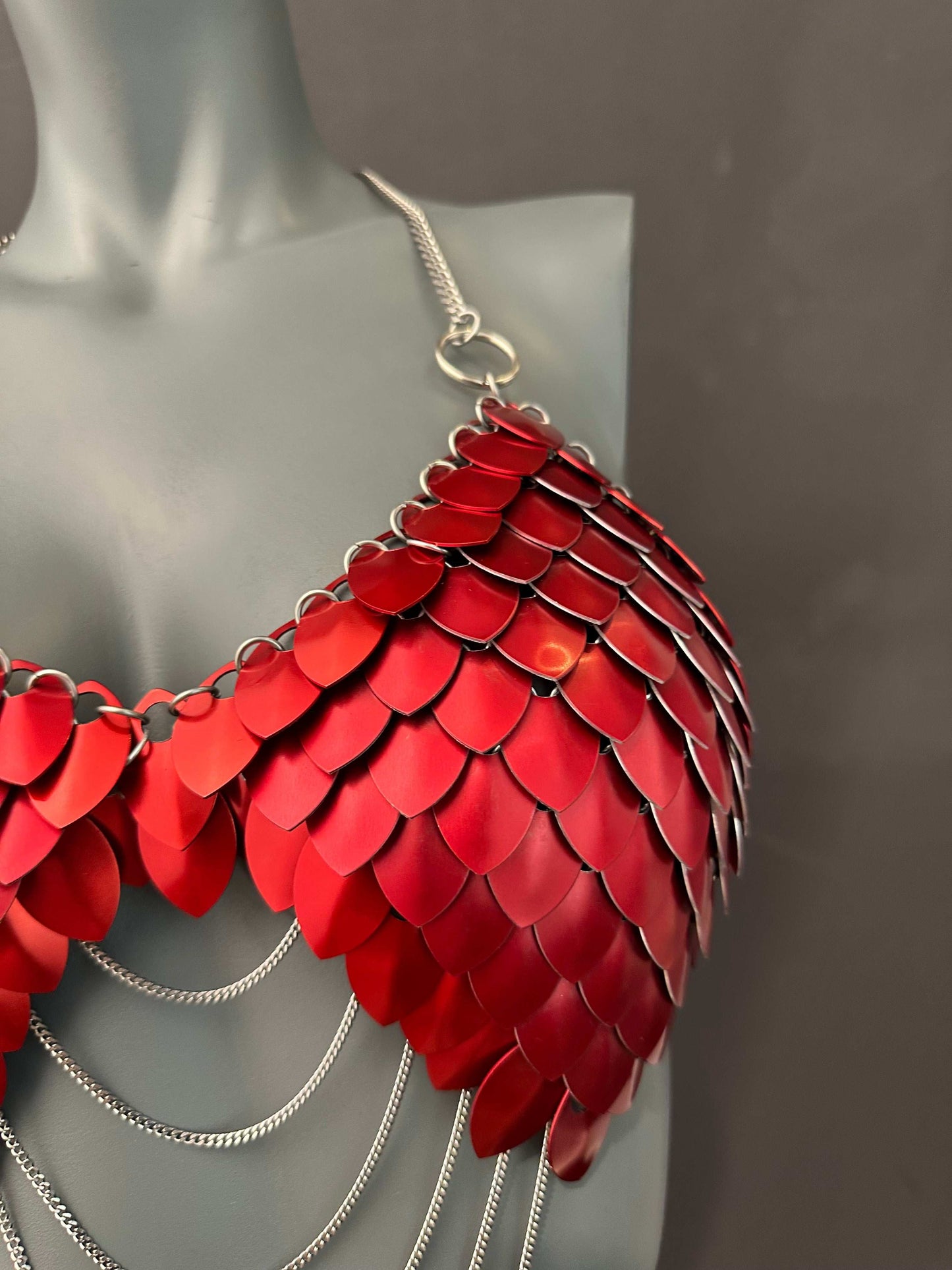 Red scale metal bra with silver chains on gray mannequin, bold and edgy fashion statement