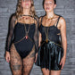 Red and gold body chain jewelry on two confident women in black lace and leather outfits against a stone wall