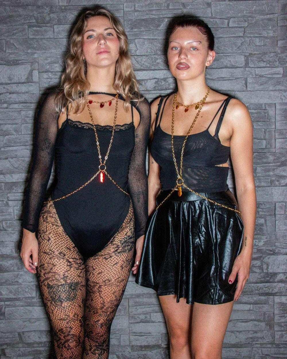 Red and gold body chain jewelry on two confident women in black lace and leather outfits against a stone wall